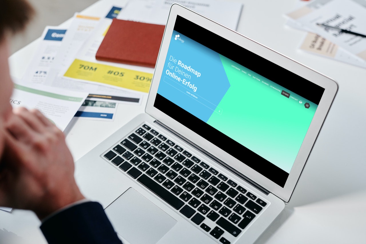 Das Bild zeigt ein 3D Mockup der Website eology.de. Das Mockup wird auf einem Laptop dargestellt. Vor dem Laptop ist verschwommen eine Person zu erkennen. Der Laptop steht auf einem Tisch. Auf dem Tisch sind noch weitere Unterlagen im Hintergrund zu sehen.